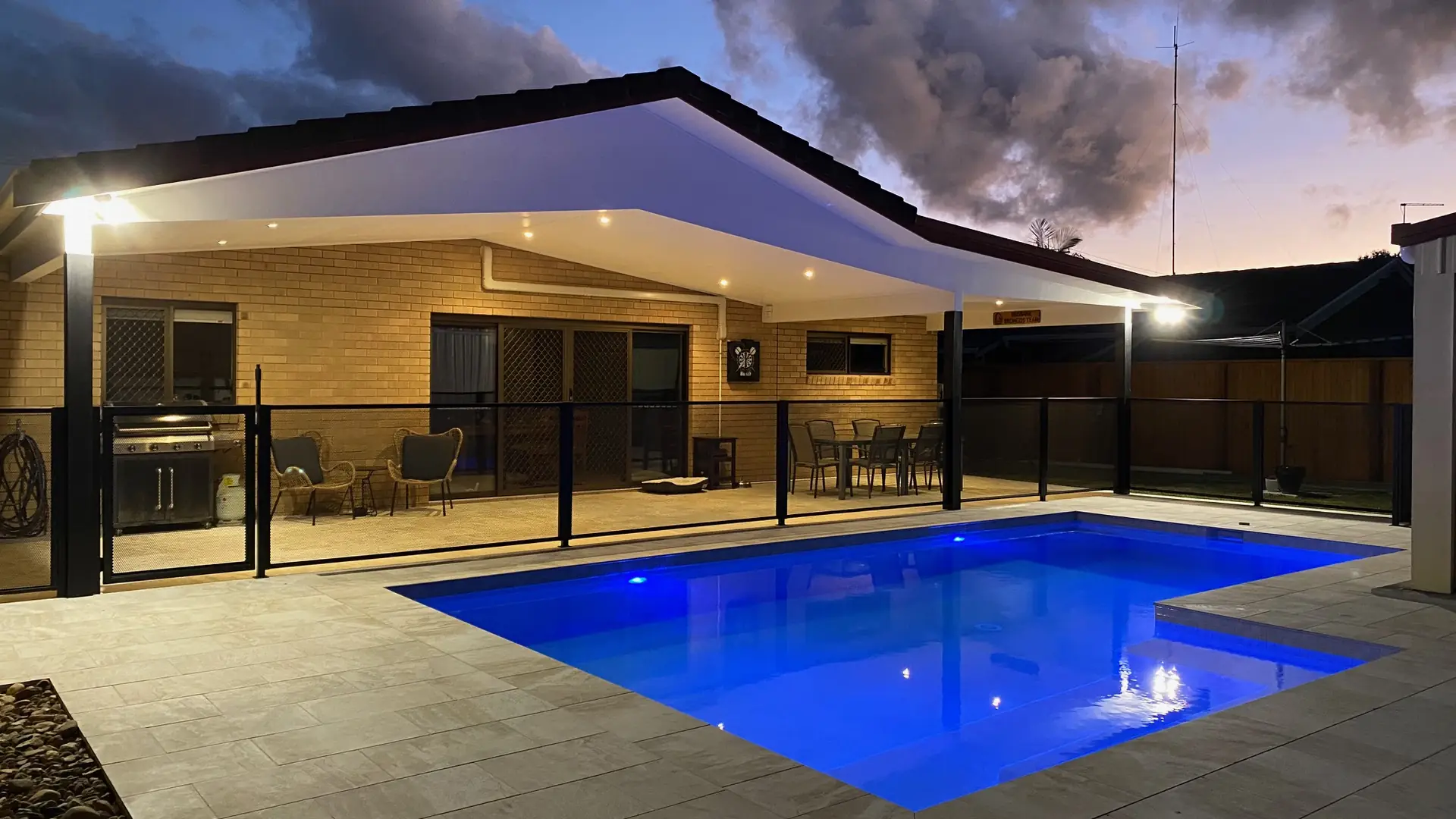 A pool glowing blue at dusk, with the patio seen straight through a black PoolPerf&reg; pool fence