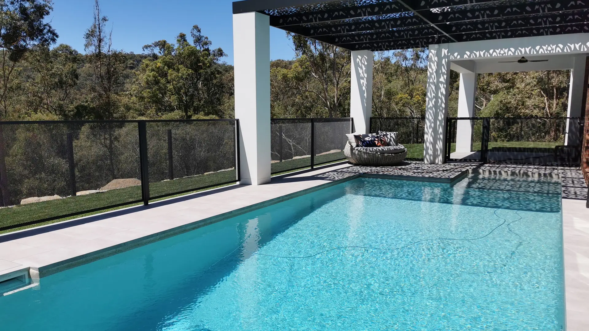 A bright blue pool beside a white pergola, with bushland showing through a PoolPerf&reg; pool fence
