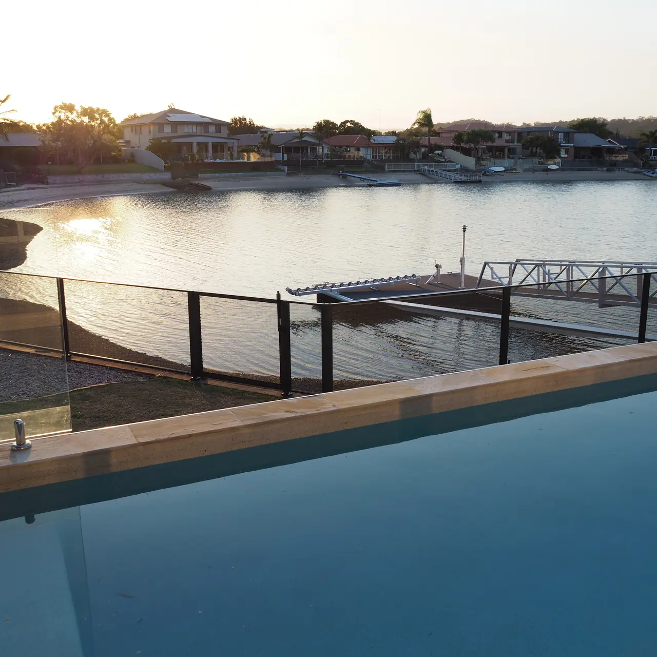 A pool in Queensland above a canal at sunset, with a black PurePerf fence and gate along the water's edge and a jetty beyond