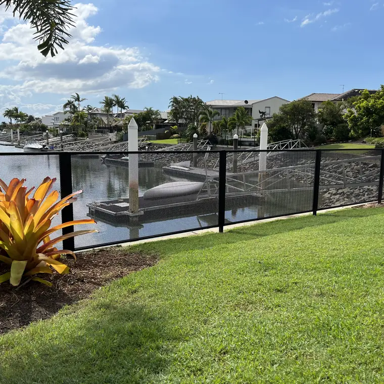 A black perforated fence along a canal in Queensland, with a pontoon, boats and canal-front homes seen through it
