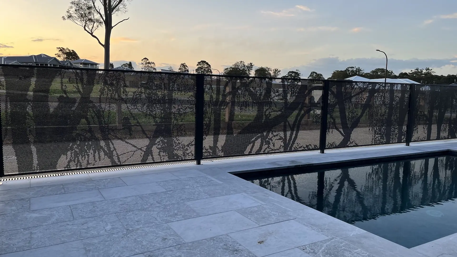 A black pool fence with gum tree silhouettes cut into it, beside a dark pool at sunset