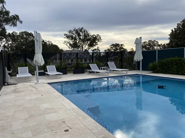 A black Gum Perf design pool fence behind white sun lounges and a pool