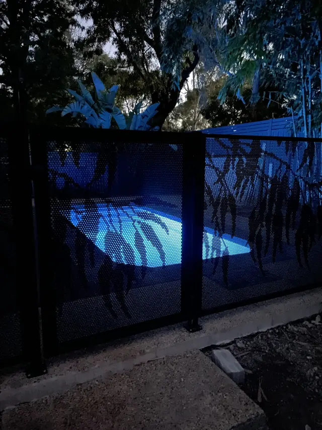 A black Gum Perf design fence at night, with the lit pool showing through