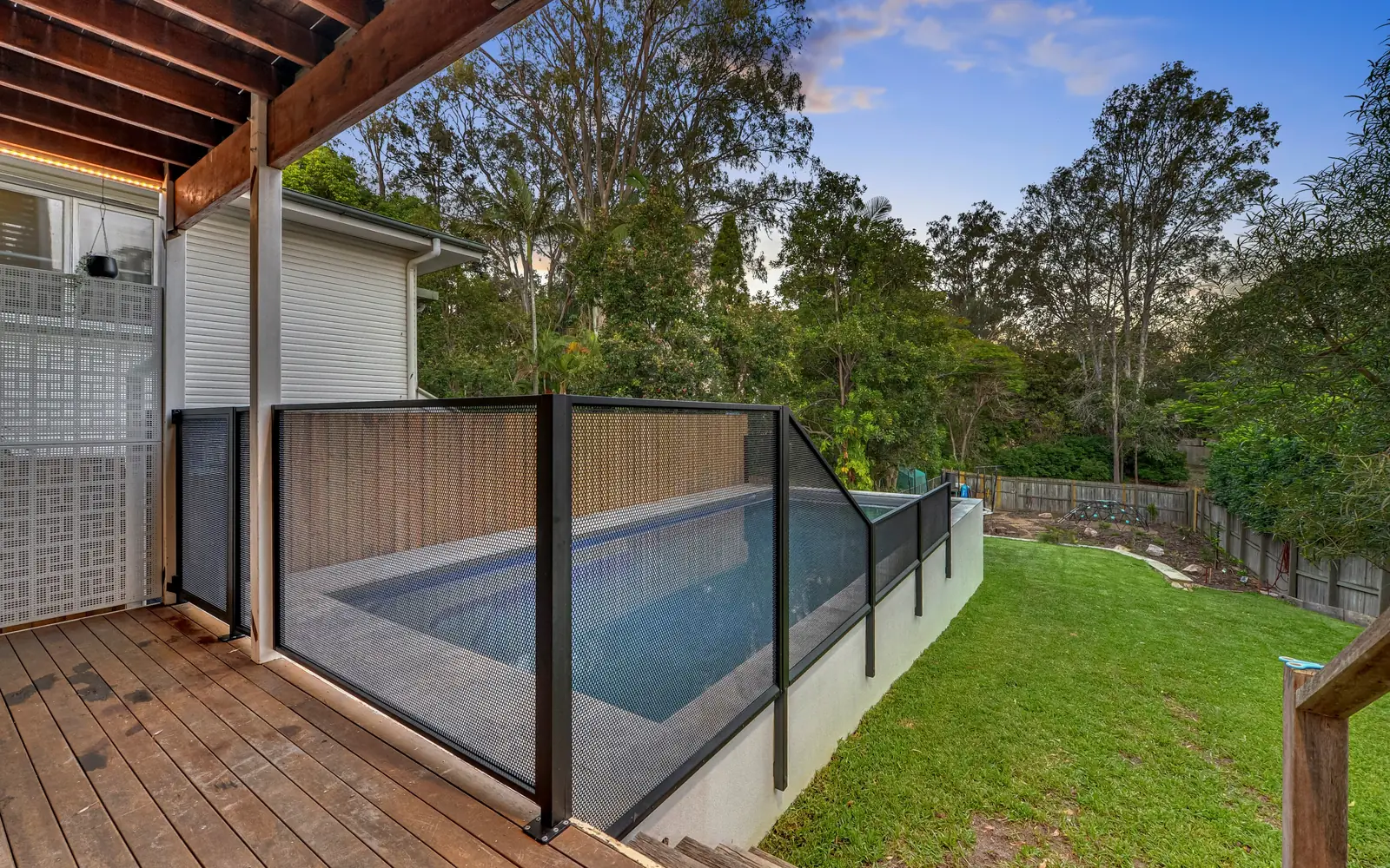 A timber deck beside a raised pool, with a dark see-through fence along the edge of the pool
