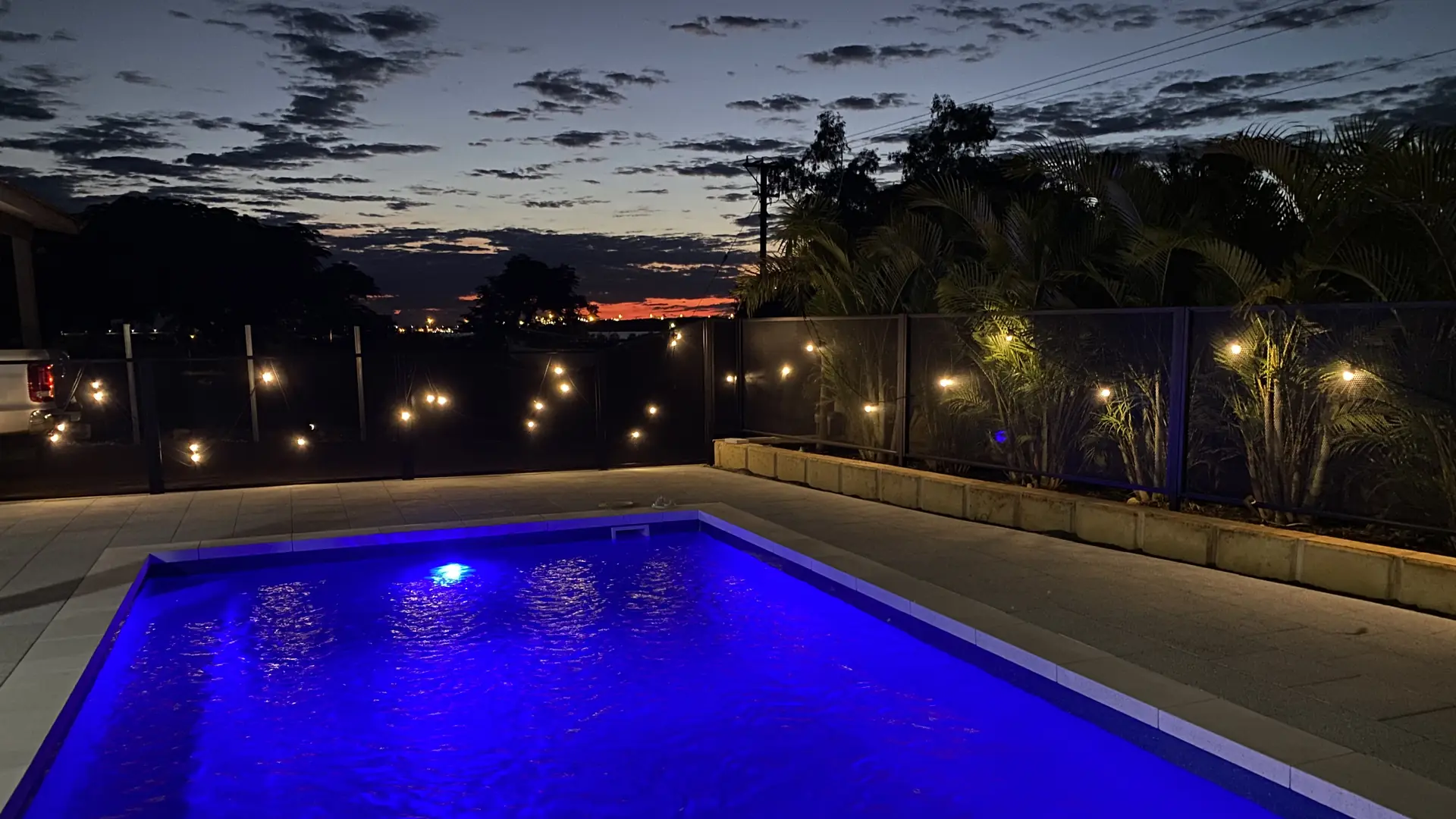A pool lit blue at dusk, with festoon lights glowing through a black pool fence