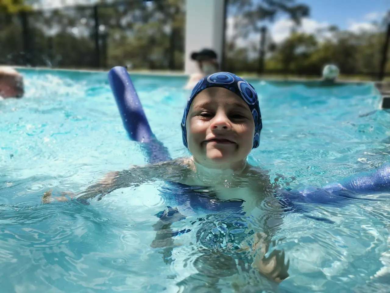 One of Jason's sons in the family pool with goggles and a pool noodle, the PoolPerf fence behind him