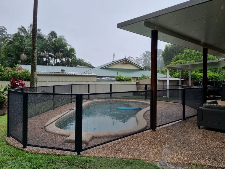 A black PoolPerf fence curving around a kidney shaped pool on pebble paving, next to a covered patio