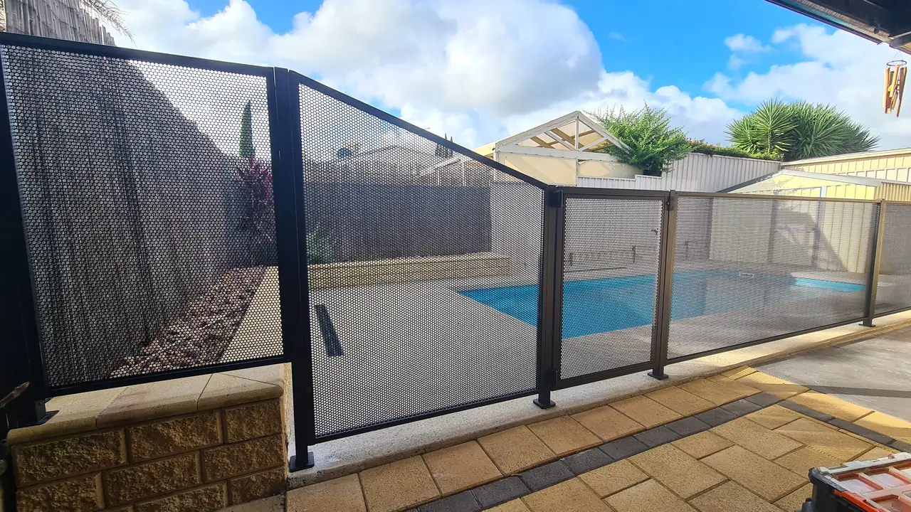 A tall black PoolPerf panel beside a raised garden bed, then a panel whose top slopes down to normal fence height at the gate, with a pool behind