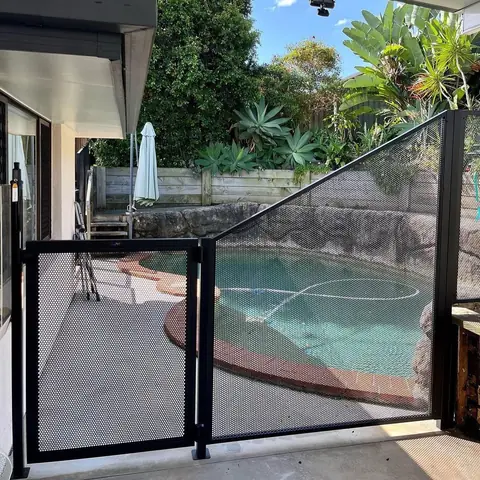 A black PoolPerf gate and a panel whose top slopes up to a tall post beside a rock garden behind a pool