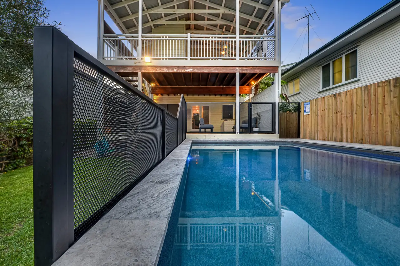 A long lit pool at dusk, with black PoolPerf panels along its raised edge and a two storey Queensland home at the far end