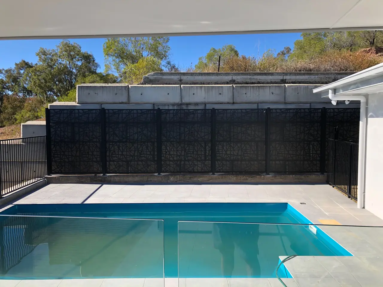A wall of black Bird Sticks design panels across the back of a pool, below a concrete retaining wall