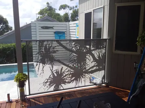 A light coloured Pandanus design panel on a timber deck beside a pool, the frond shadows across it