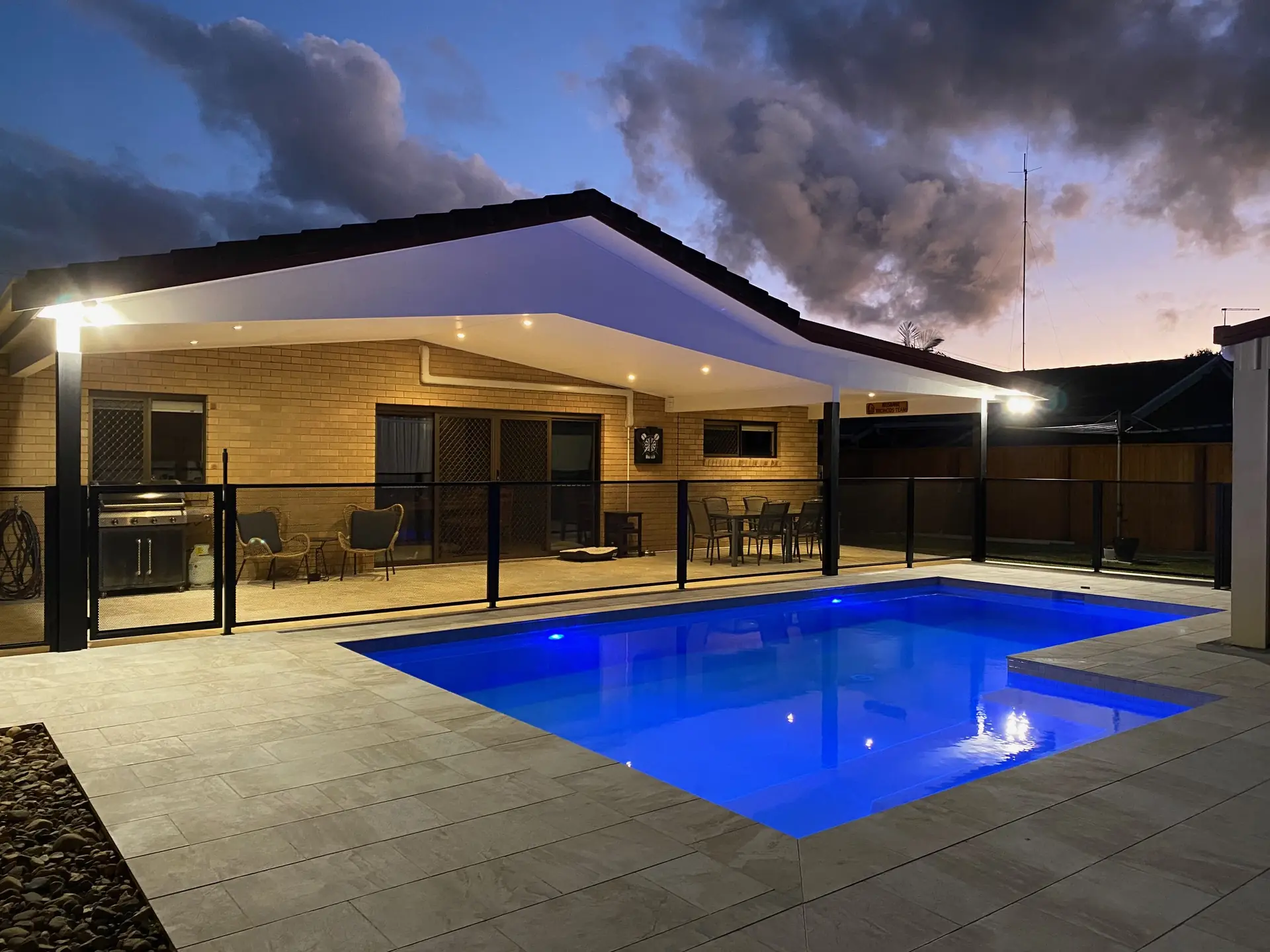 A black PurePerf fence around a lit pool at dusk in Queensland, with the covered outdoor room glowing behind it