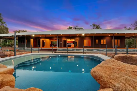 A rock-edged pool in front of a long lit home at dusk, with silver Wave design panels along the far side