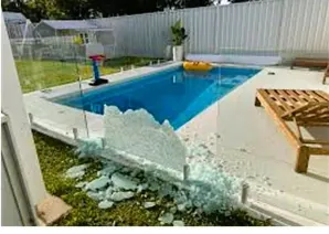 A shattered glass pool fence panel in pieces on the grass beside a pool after an extreme change in temperature
