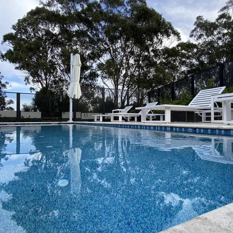 A large tiled pool with sun lounges and a black PoolPerf fence at the far end, under gum trees