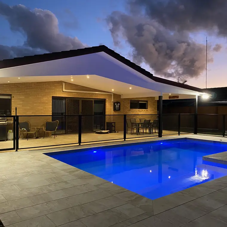 A lit blue pool at dusk with a black PoolPerf fence between it and the covered outdoor room behind