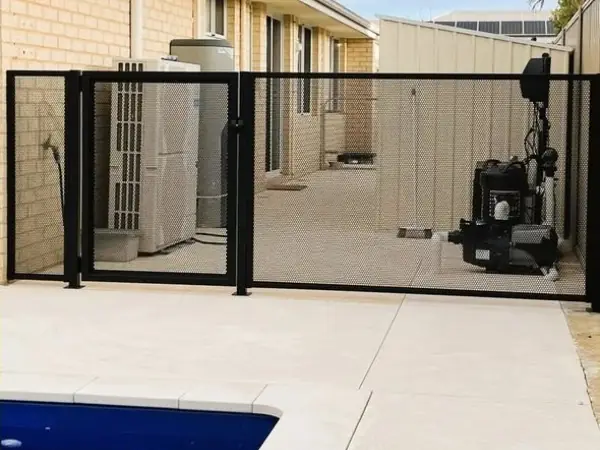 A black perforated fence and wide gate hiding an air conditioning unit and a water tank beside a house