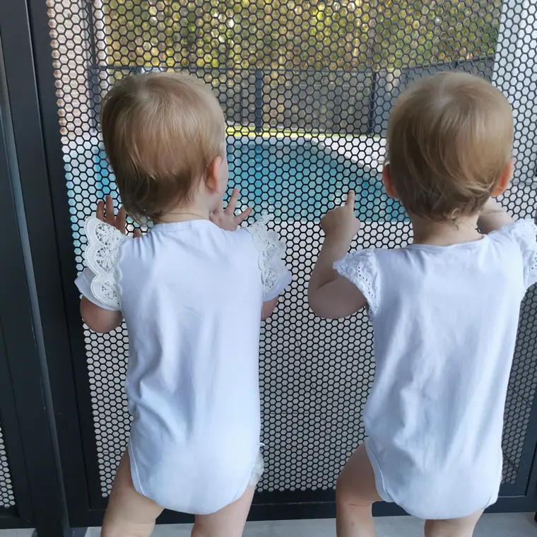 Two toddlers from behind, hands on a PoolPerf panel, the pool visible through the holes