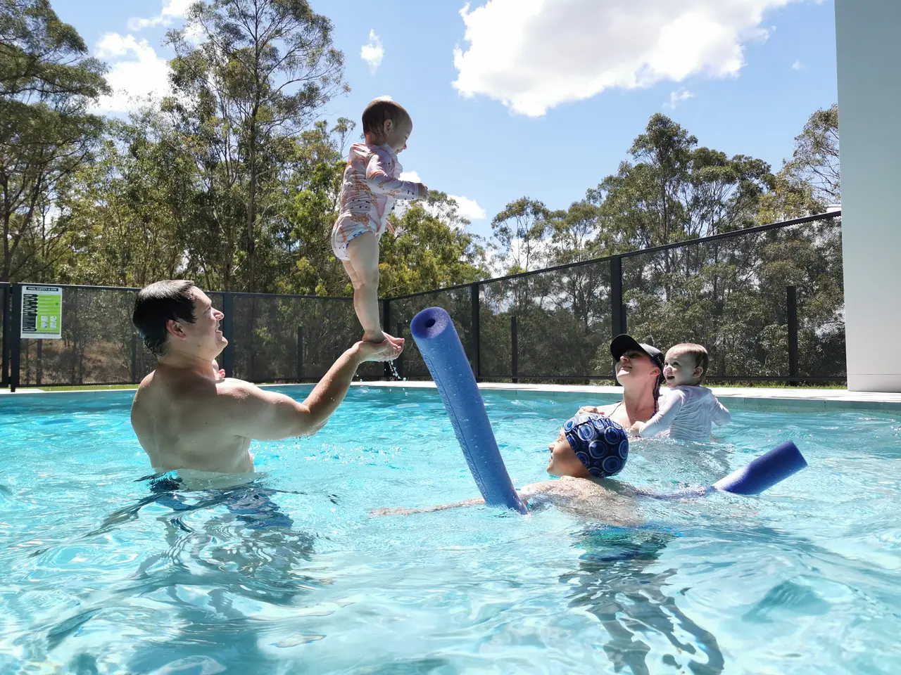 A dad in the pool lifting his toddler into the air while mum, a baby and an older child play nearby, with a PoolPerf fence behind them