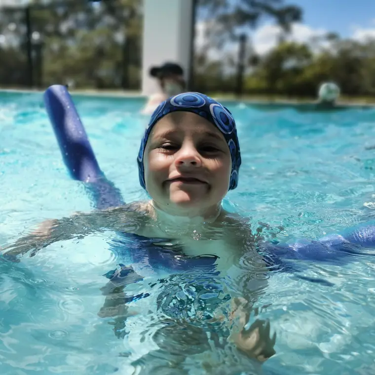 A smiling boy in goggles floating on a pool noodle in the family pool