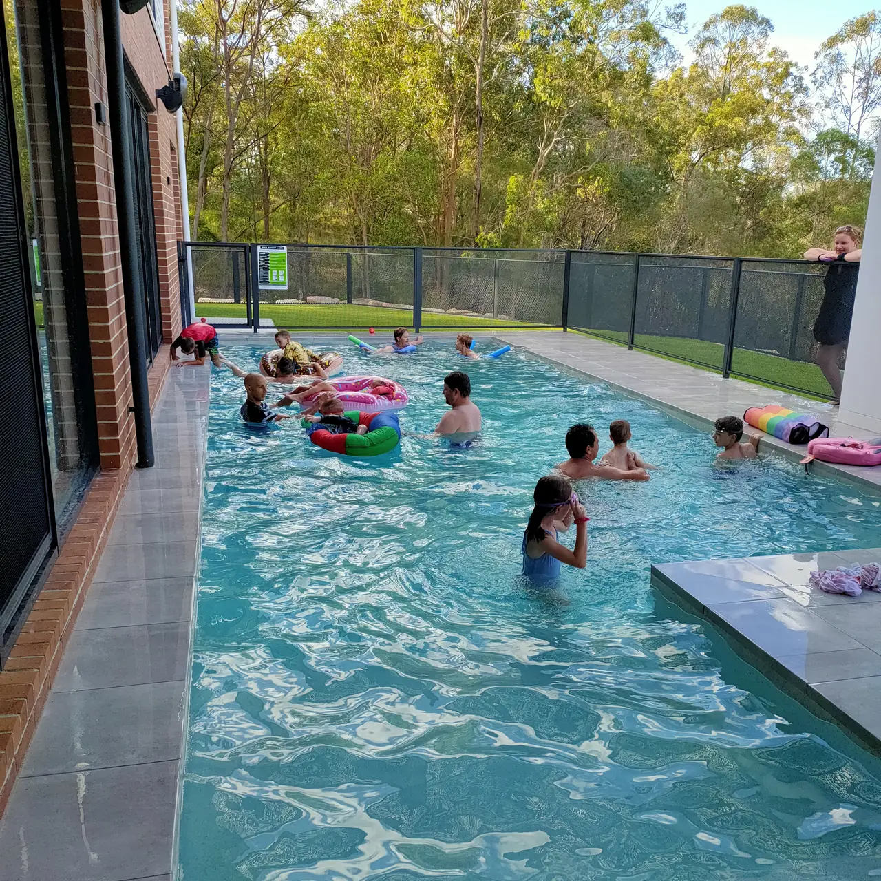 A long pool full of children swimming, with a PoolPerf fence running along the yard behind them