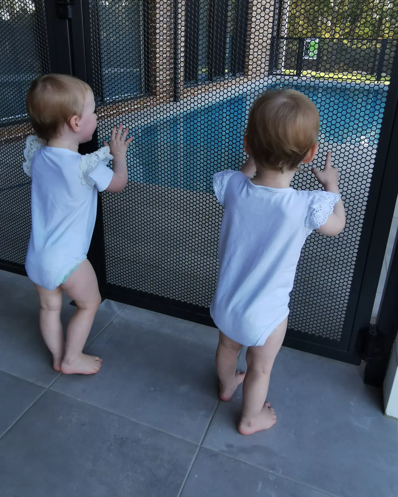 Twin toddlers standing at a PoolPerf pool fence with their hands on the panel, looking through it at the pool