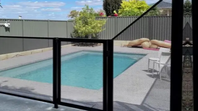 A black PoolPerf fence and gate beside a pool, with one panel rising on an angle to a taller post