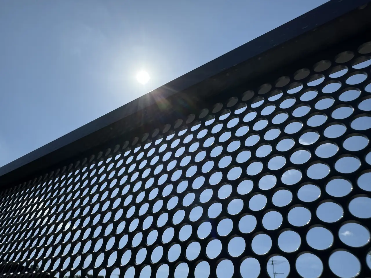 A PoolPerf PurePerf panel photographed from below against a blue sky, with the sun shining over the top rail and the sky showing through every round hole