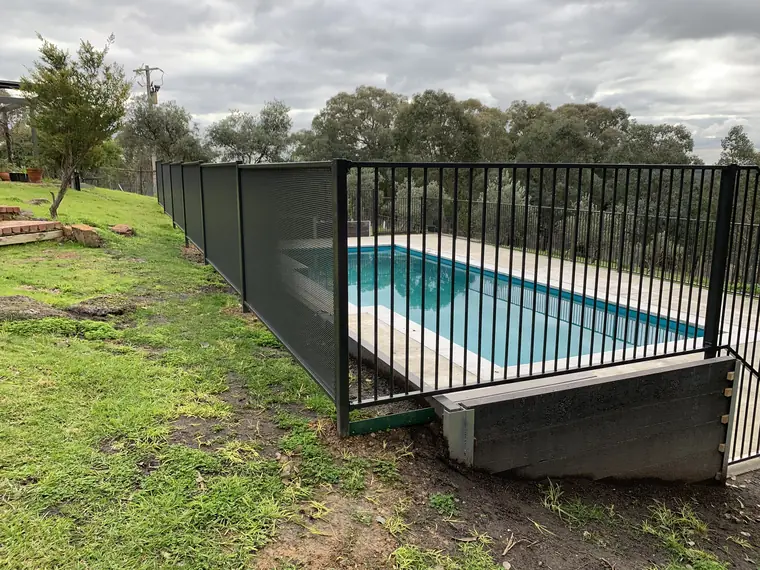A pool on a sloping lawn in Victoria fenced two ways: black PoolPerf panels along the left side, and a flat-top fence of upright bars across the front