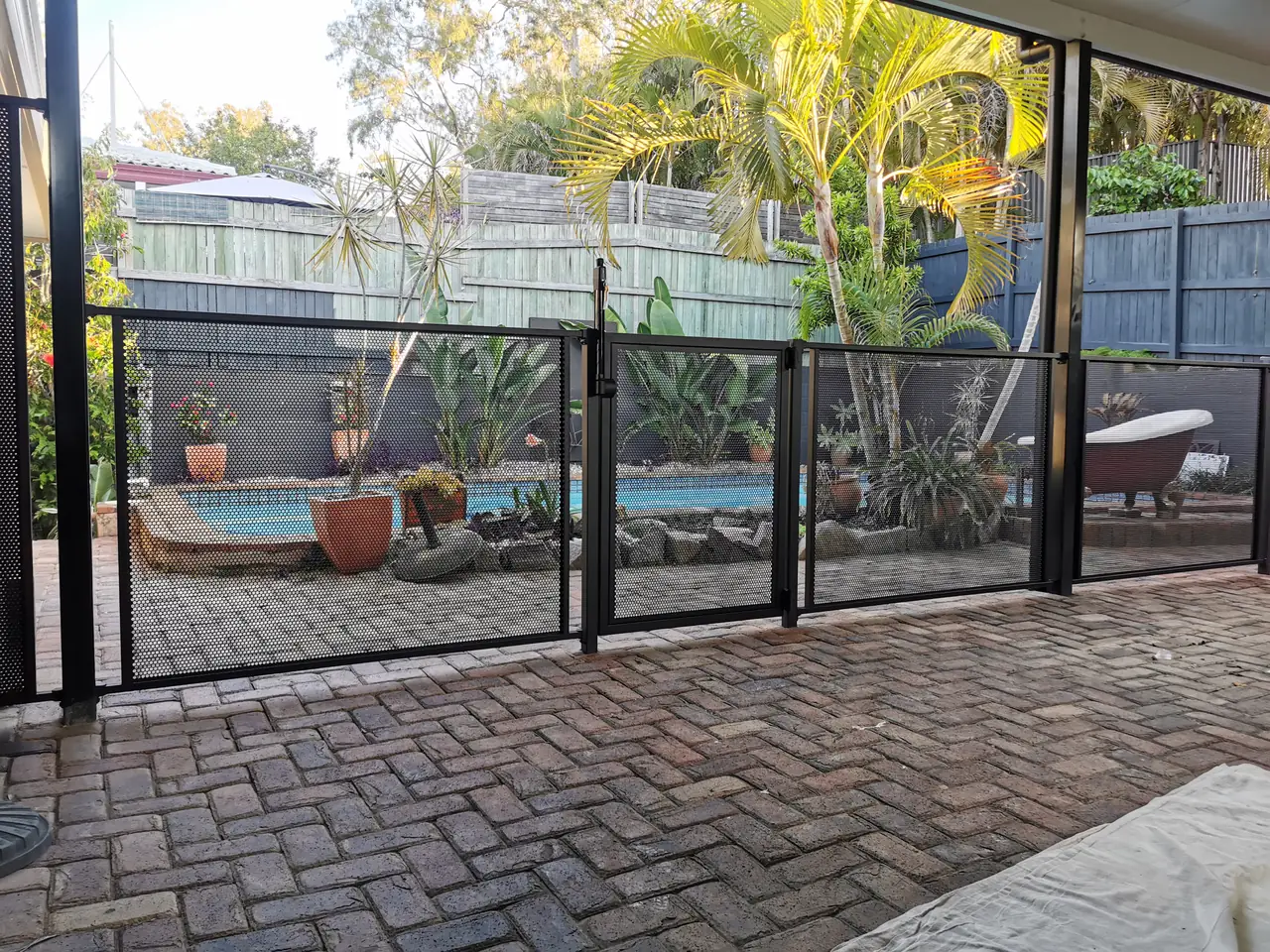 A black PurePerf pool gate in a plain black PurePerf fence, seen from a paved patio, with the pool and garden behind
