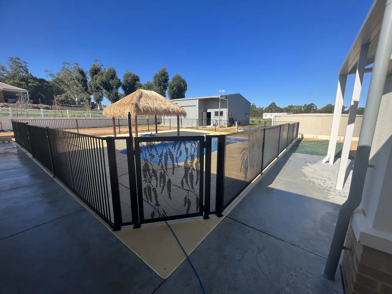 A black Gum design PoolPerf gate made to fit a black flat top pool fence, beside a pool with a thatched umbrella
