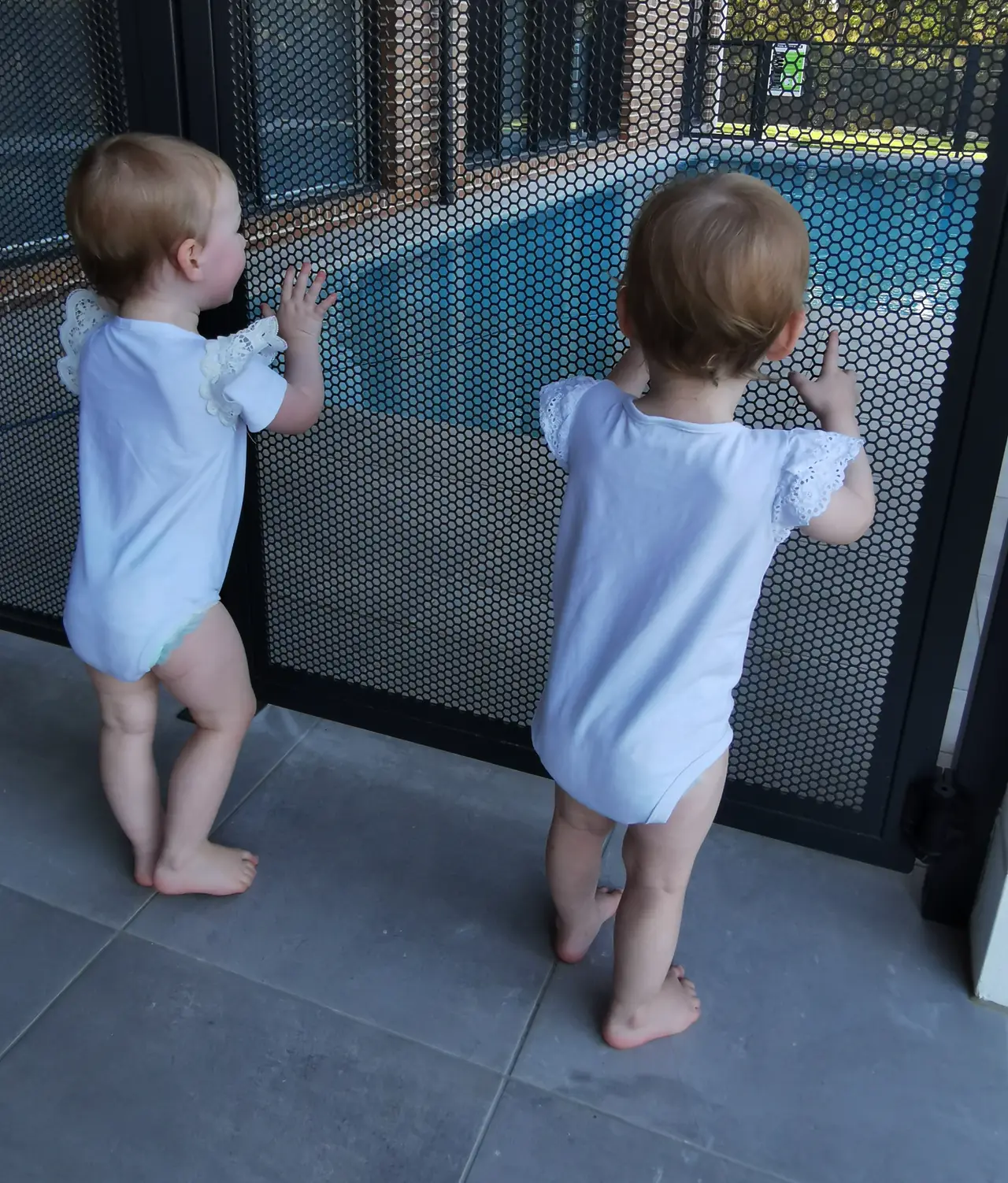 Twin toddlers in white standing at a PurePerf pool fence, looking through it at the pool