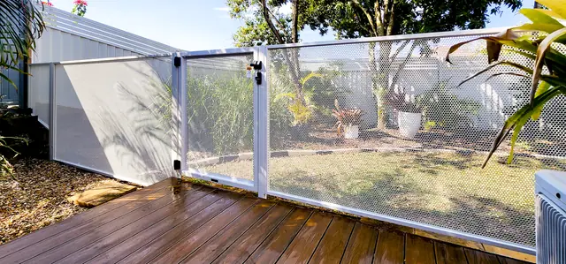 A white Pure Perf pool fence and gate between a timber deck and a tropical garden