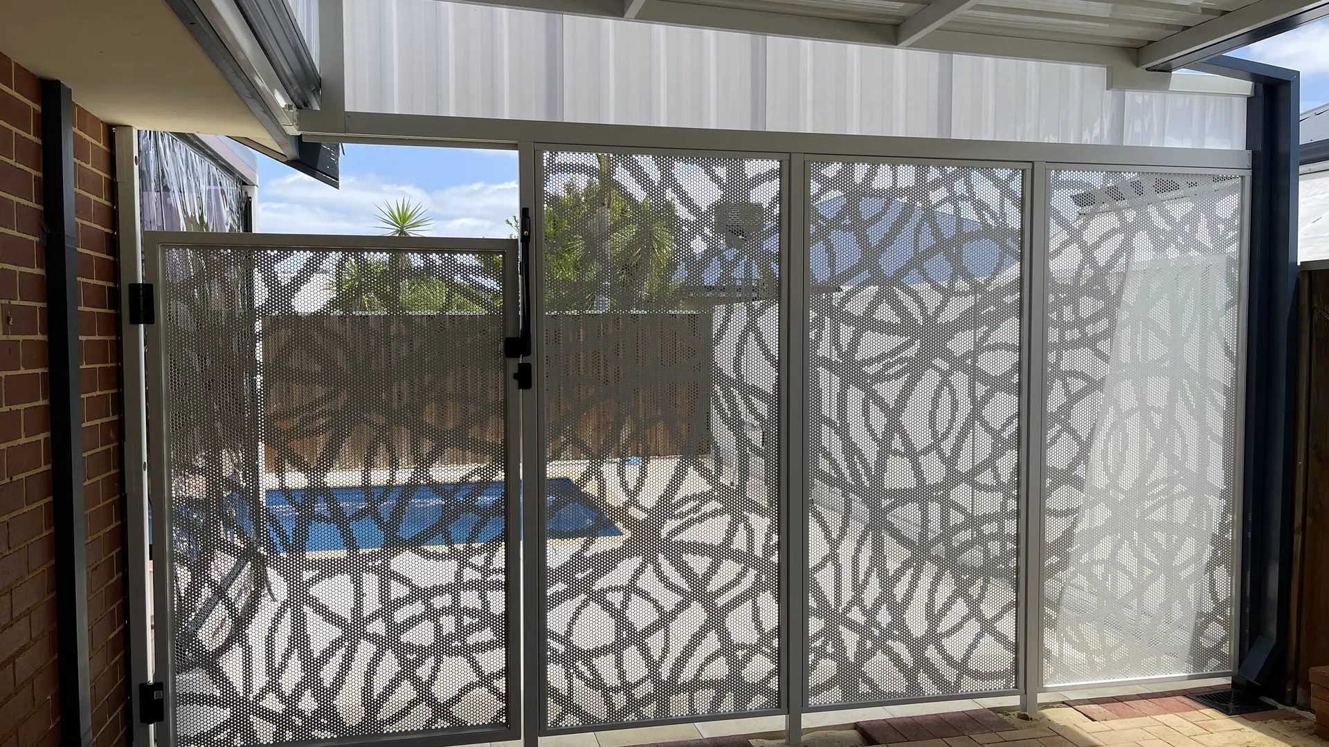 A white patterned pool gate and fence under a patio, the pool visible through the pattern