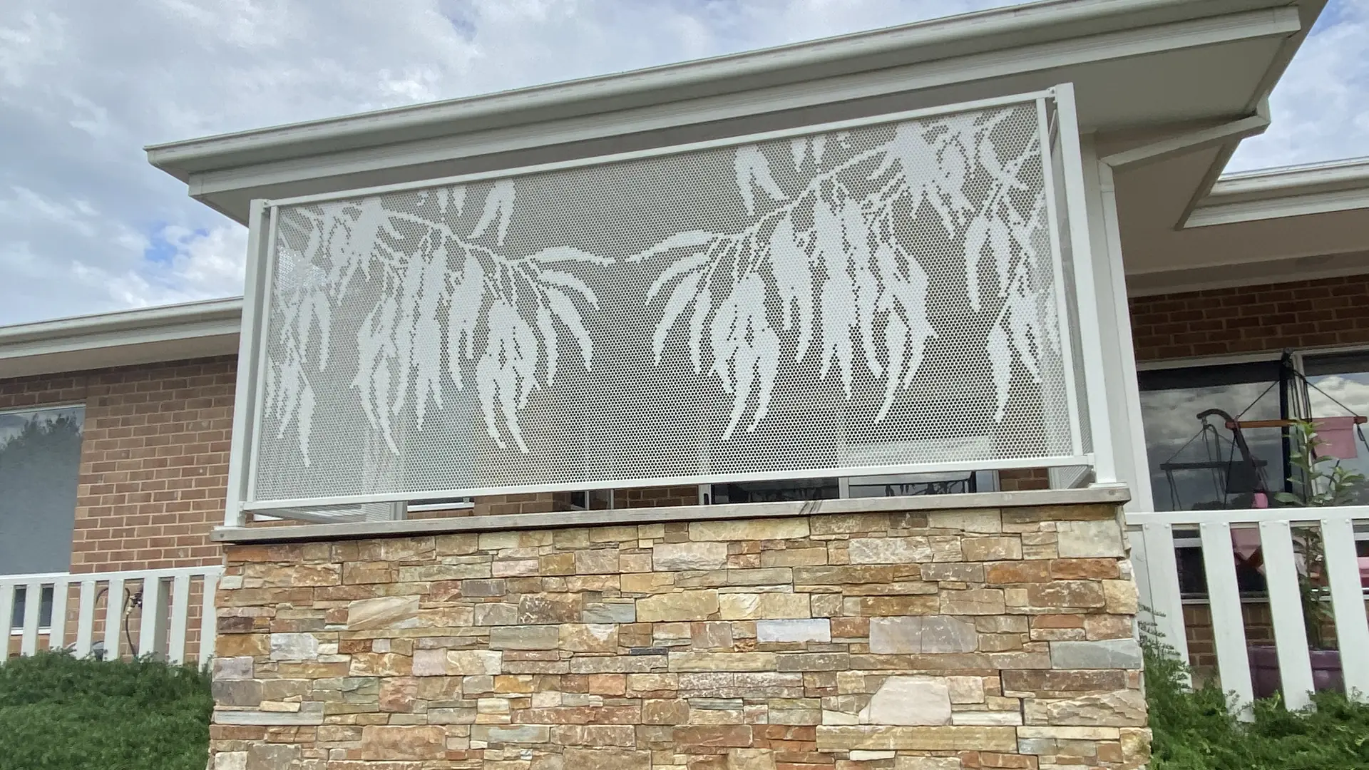 A white Gum design panel on a stone wall, with a white picket fence and white window trims beside it