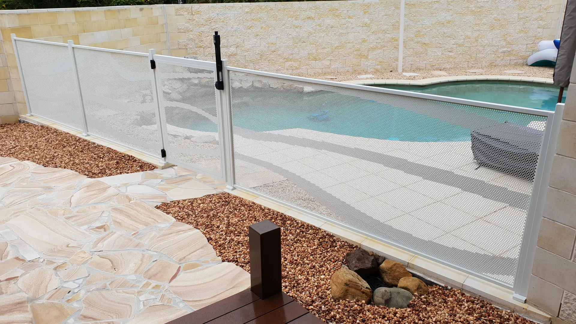 A pale Pure Perf pool fence around a freeform pool with sandstone paving and a garden bed